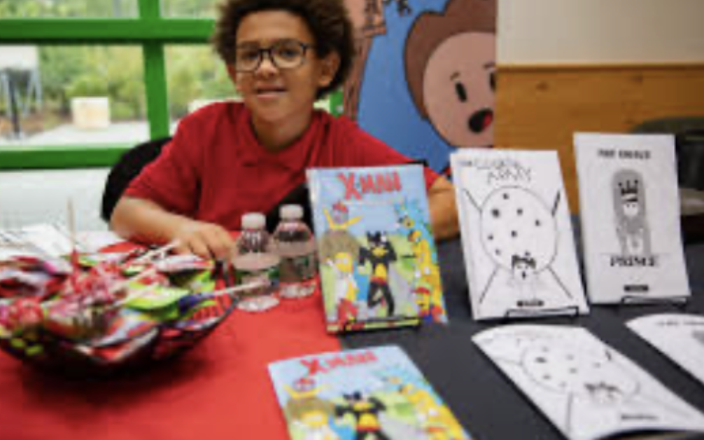 A person smiles behind a table displaying books and drawings at an event.
