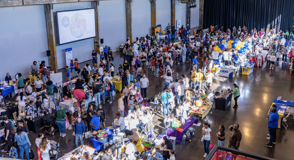 A crowded event hall with many people and tables filled with items.