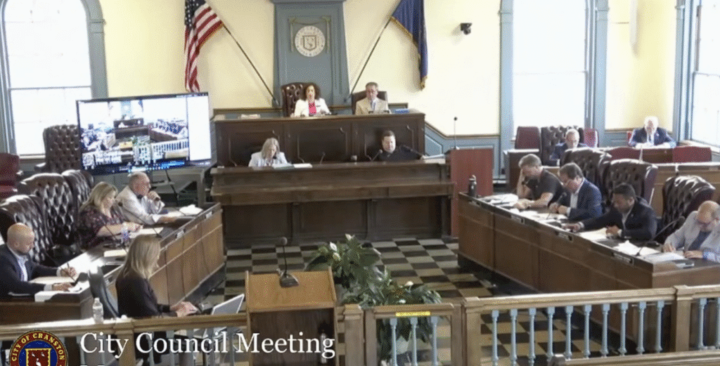 City council meeting in a formal chamber with members seated and American flags displayed.