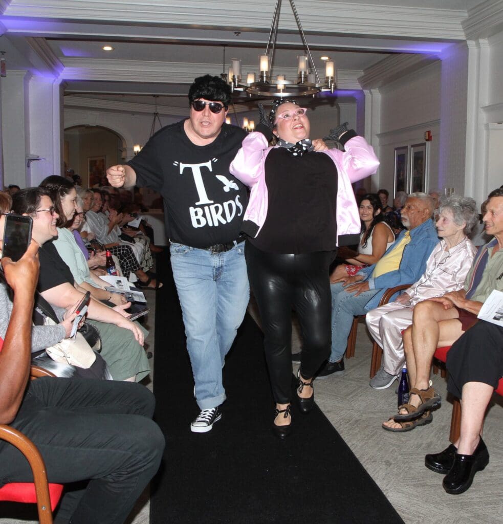 Two men walk down a runway in a fashion event with seated audience.