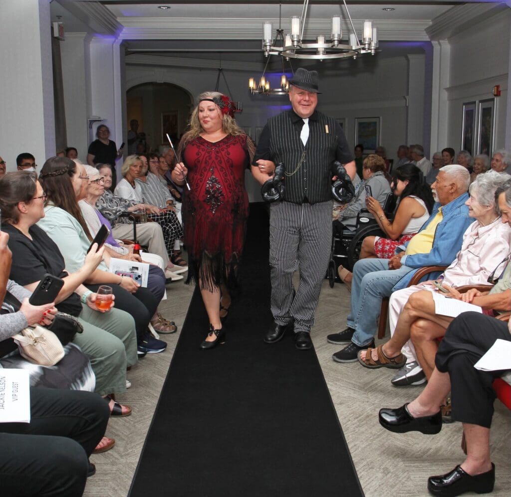A couple walks down a runway during a fashion show with seated audience.