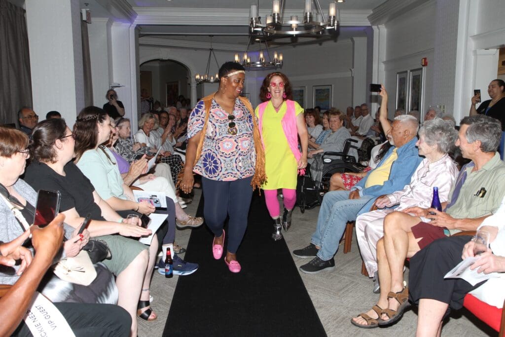 Two women walking down a runway at a fashion show with an audience seated on both sides.