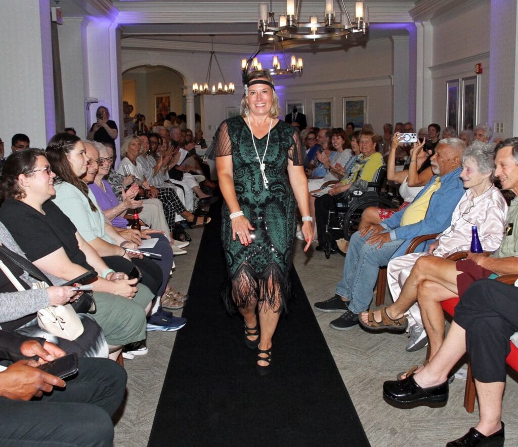 Model walks down runway in a stylish black dress at a fashion show.
