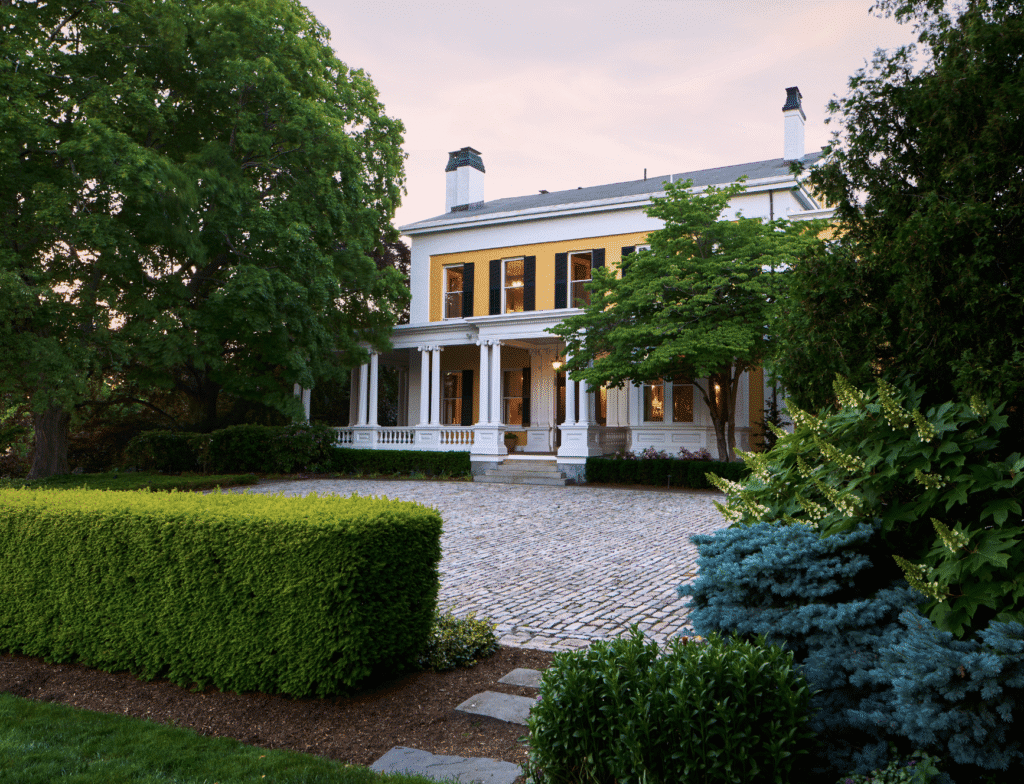 Elegant white mansion with columns surrounded by lush greenery and a cobblestone driveway.