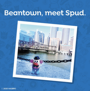 Spud toy posing by Boston's waterfront with city skyline background.