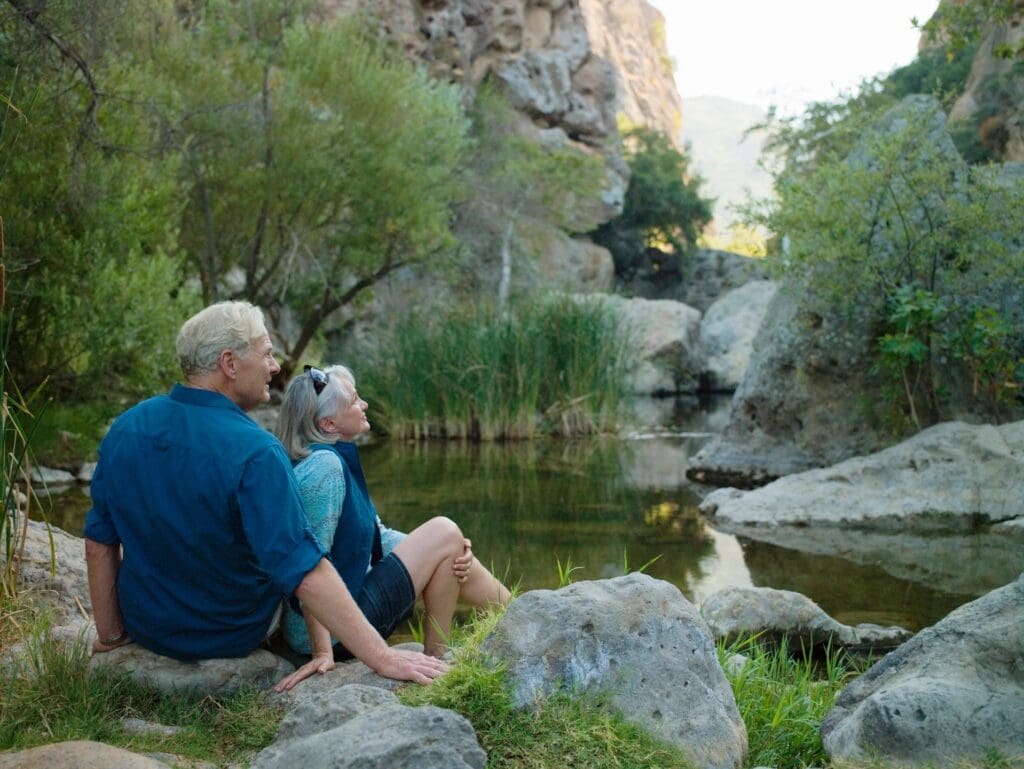 Elderly couple sitting by a serene pond surrounded by rocks and greenery.