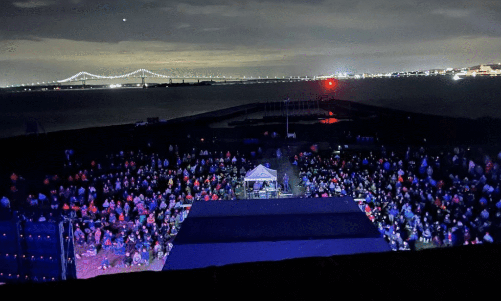 Large crowd gathered at an outdoor nighttime event with city lights in the background.