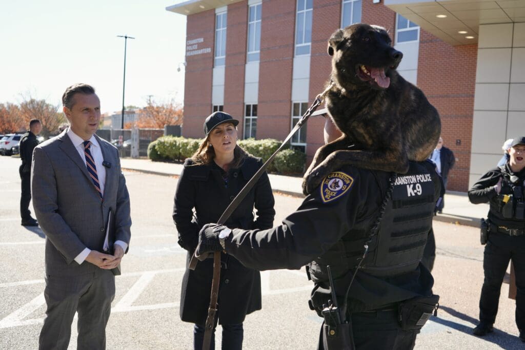 A police officer with a police dog talking to two civilians outside.