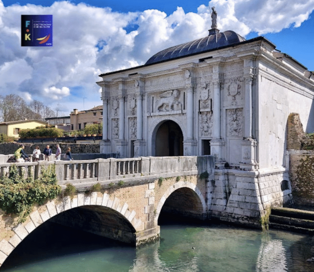 Historic white stone bridge and gate over calm river under blue sky.
