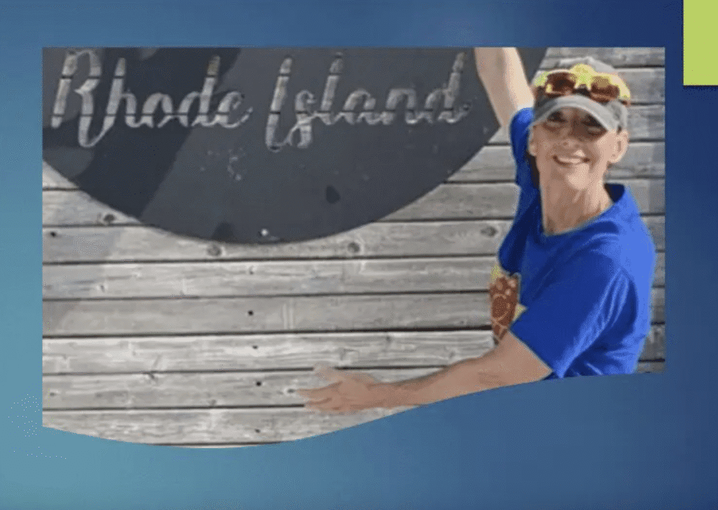 Smiling man posing by a wooden sign that reads 'Rhode Island'.