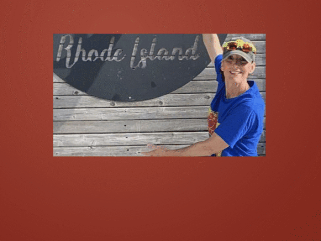 A man in sunglasses and a cap pointing at a Rhode Island sign.
