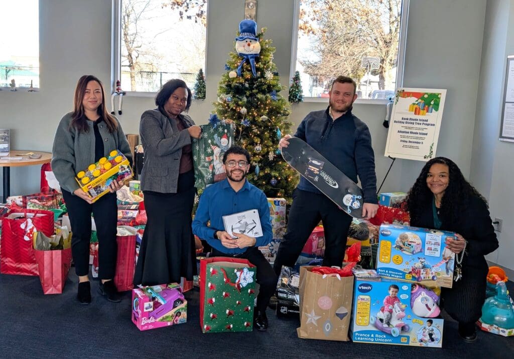 Group of four people posing with wrapped holiday gifts and a decorated Christmas tree indoors.