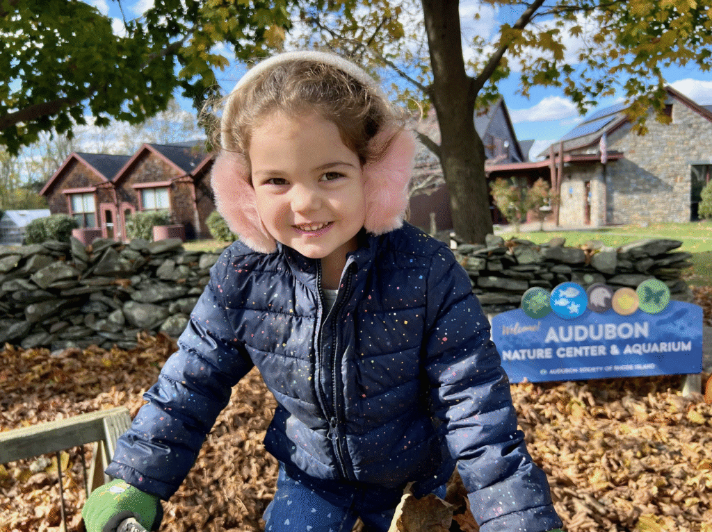 Smiling toddler in a blue jacket playing outdoors on a sunny day.