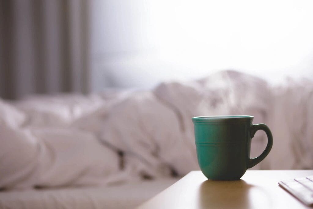 A green mug on a wooden surface with a blurred white bed in the background.