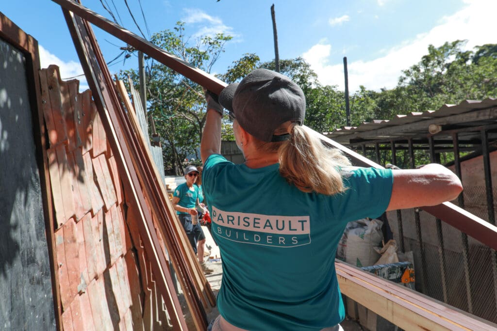 Person wearing a turquoise shirt and cap working outdoors with wooden materials.