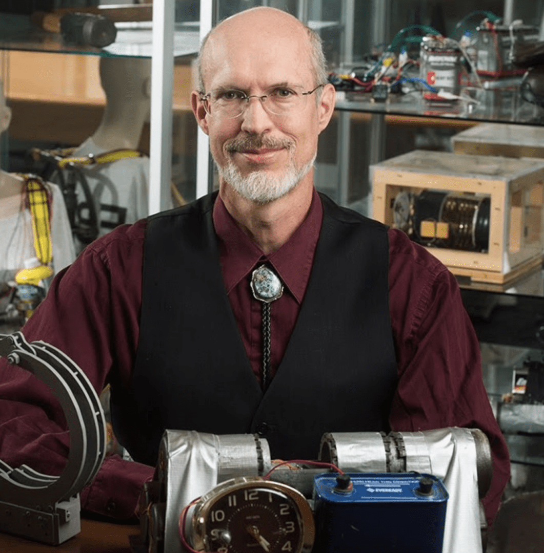 An older man in a vest and bolo tie in a workshop with mechanical devices.
