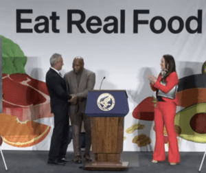 Three people on stage at an event with 'Eat Real Food' banner.