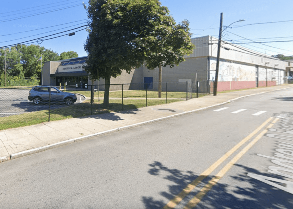Street view of a fenced industrial building with parked cars and trees.