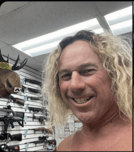 Man smiling in front of a wall of rifles and a mounted deer head.