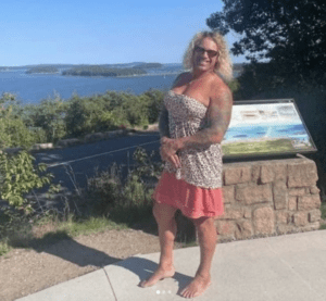 Woman posing near a scenic waterfront with greenery and blue water in the background.