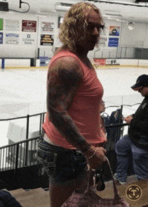 Tattooed man in a red sleeveless shirt at an ice rink.