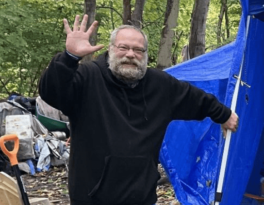 Man in black hoodie waving outdoors near a blue tarp and campsite.