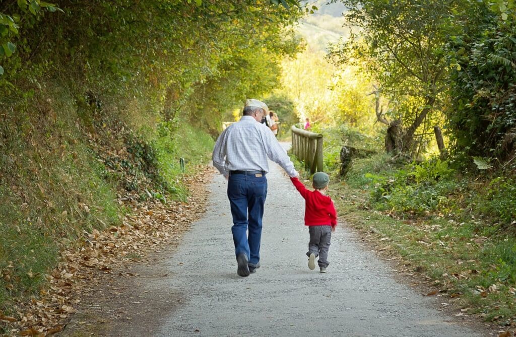 An elderly man walks hand-in-hand with a child on a tree-lined path.