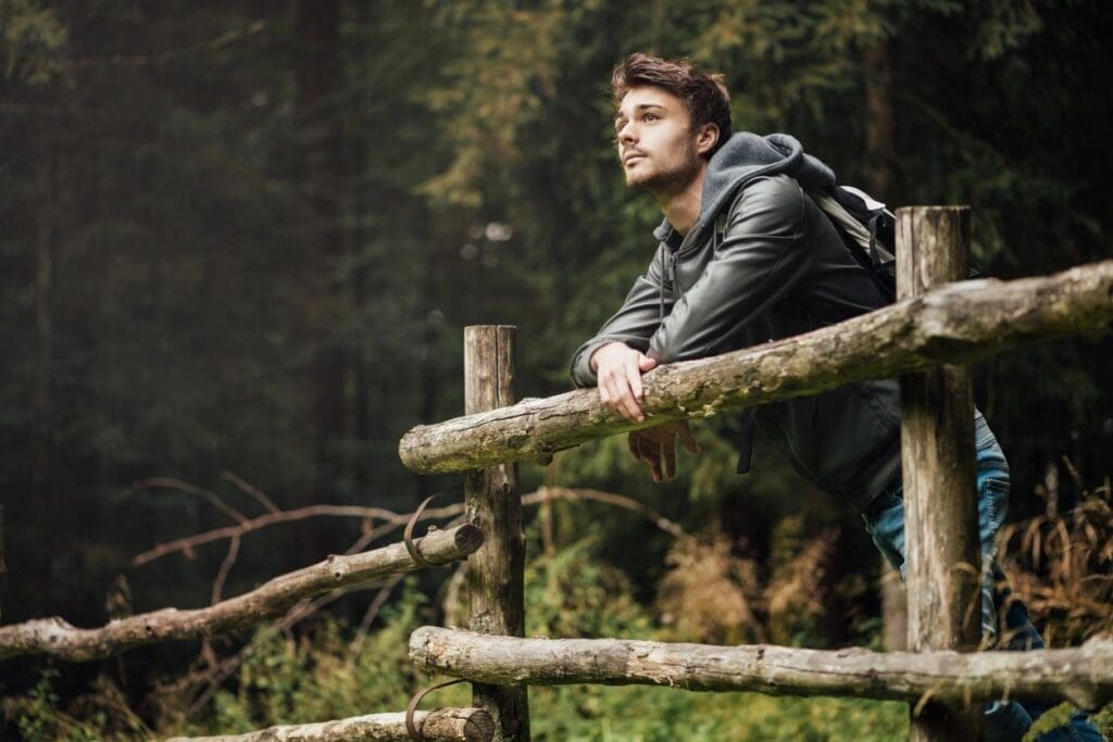Man leaning on wooden fence in a forest, looking thoughtful.