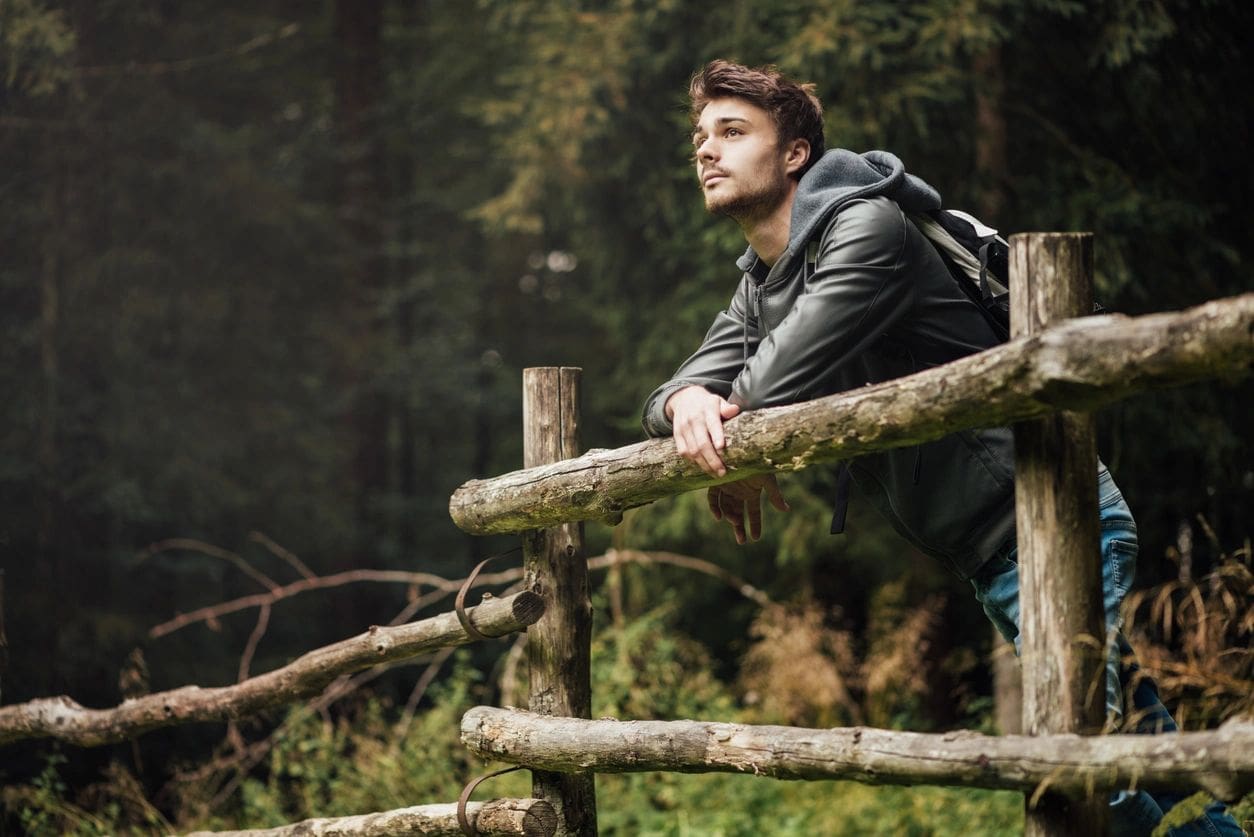 Man leaning on wooden fence in a forest, looking thoughtful.