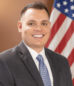 Professional headshot of a smiling man in a suit with an American flag background.