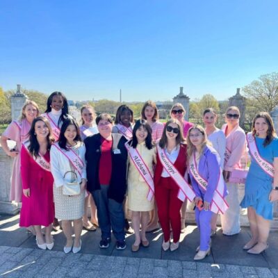 A group of women in pink sash posing for a photo under cherry blossoms.