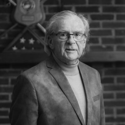 A black and white photo of an older man standing in front of a brick wall.
