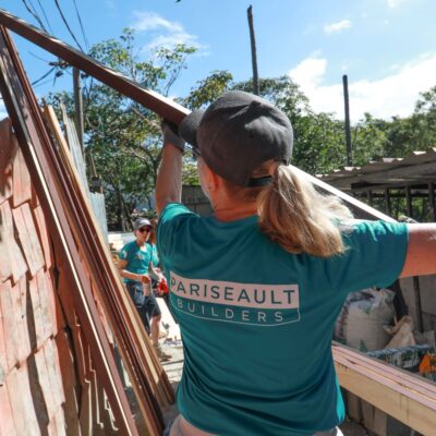 Person wearing a turquoise shirt and cap working outdoors with wooden materials.