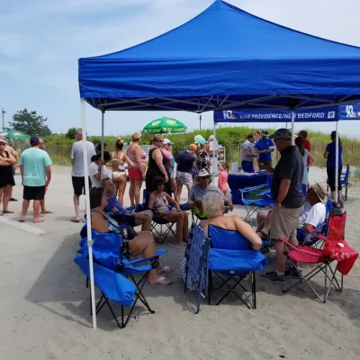A group of people sitting under a blue tent on the beach.