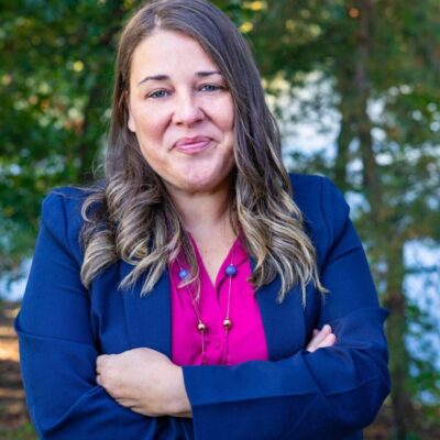 A woman in a blue blazer and pink shirt standing in front of a lake.
