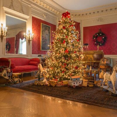 A Christmas tree adorned with sparkling lights and ornaments stands tall in a room with vibrant red walls. The festive atmosphere exudes the charm of Newport mansions during the Christmas season.
