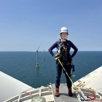 Worker in safety gear standing on a wind turbine platform overlooking the ocean.