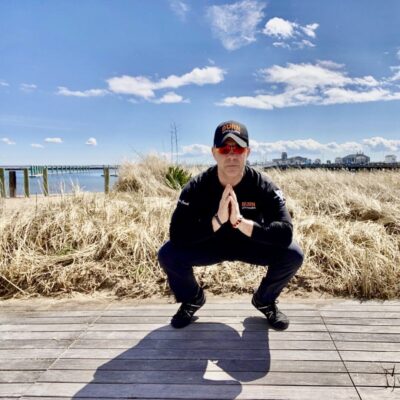 A man squatting on a boardwalk near the ocean.
