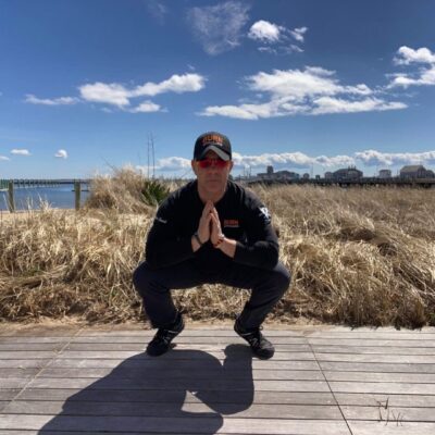 A man squatting on a boardwalk near the ocean.