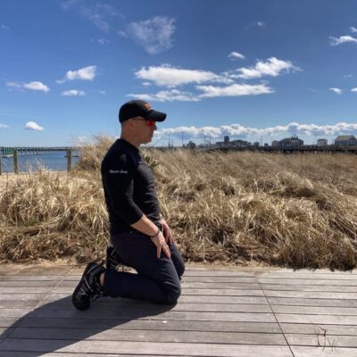 A man exercising, kneeling on a boardwalk next to a body of water.