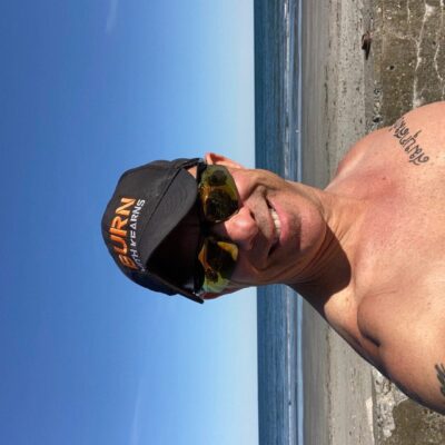 A man in a shirt and hat taking a selfie on the beach.