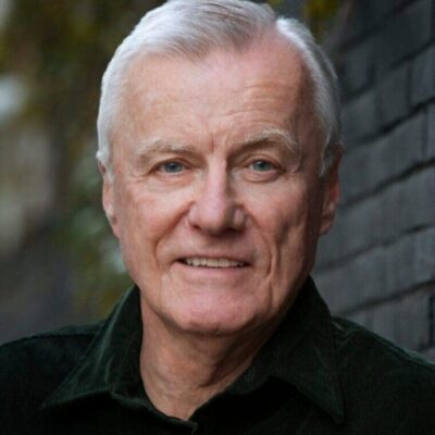 An older man smiling in front of a brick wall.