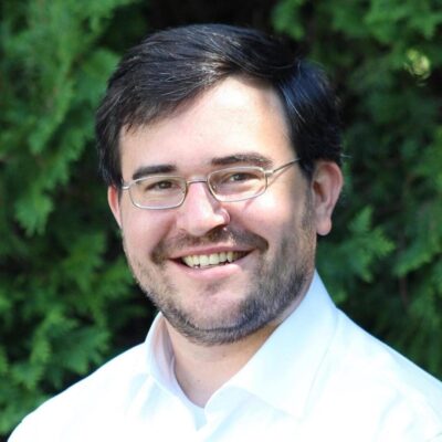 A man with glasses smiling in front of bushes.