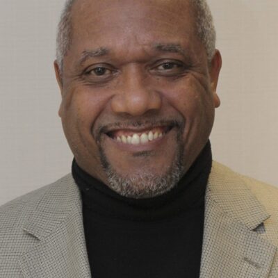 A man in a gray suit and black turtleneck smiles.