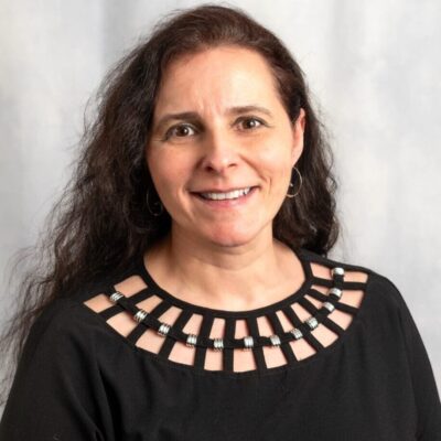 Smiling woman with long curly hair wearing a black top with a decorative neckline.