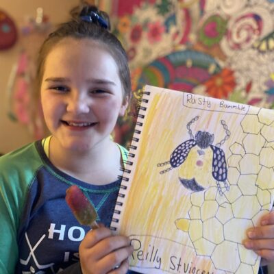 A girl holding up a notebook with a drawing of a bee.
