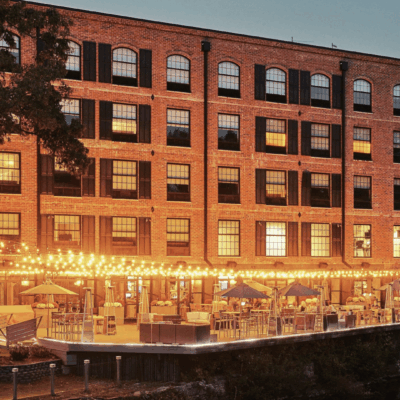 A brick building is lit up at dusk next to a river.