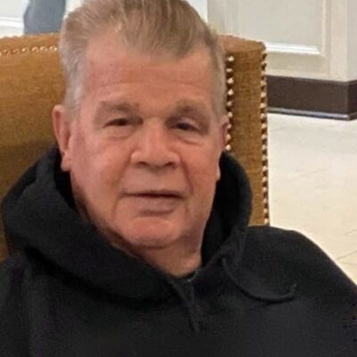 An older man in a black hoodie sitting in a chair.