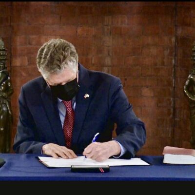 A man wearing a mask signing papers at a desk.