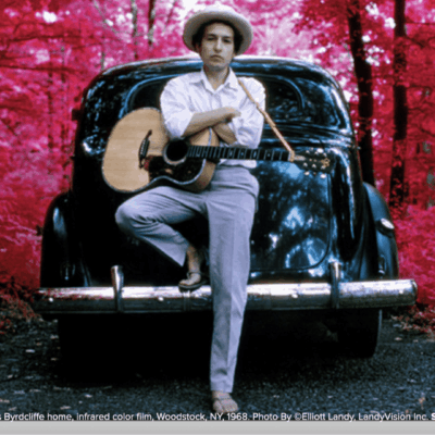 A man with a guitar leaning on the back of a car.